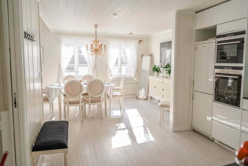 Bildet viser lit av kjøkken og stue i Holbergsuiten. Viser ovn og kjøleskap på kjøkken og spisebord med 6 stoler i stua. Lysekrone og speil i hjørnet.